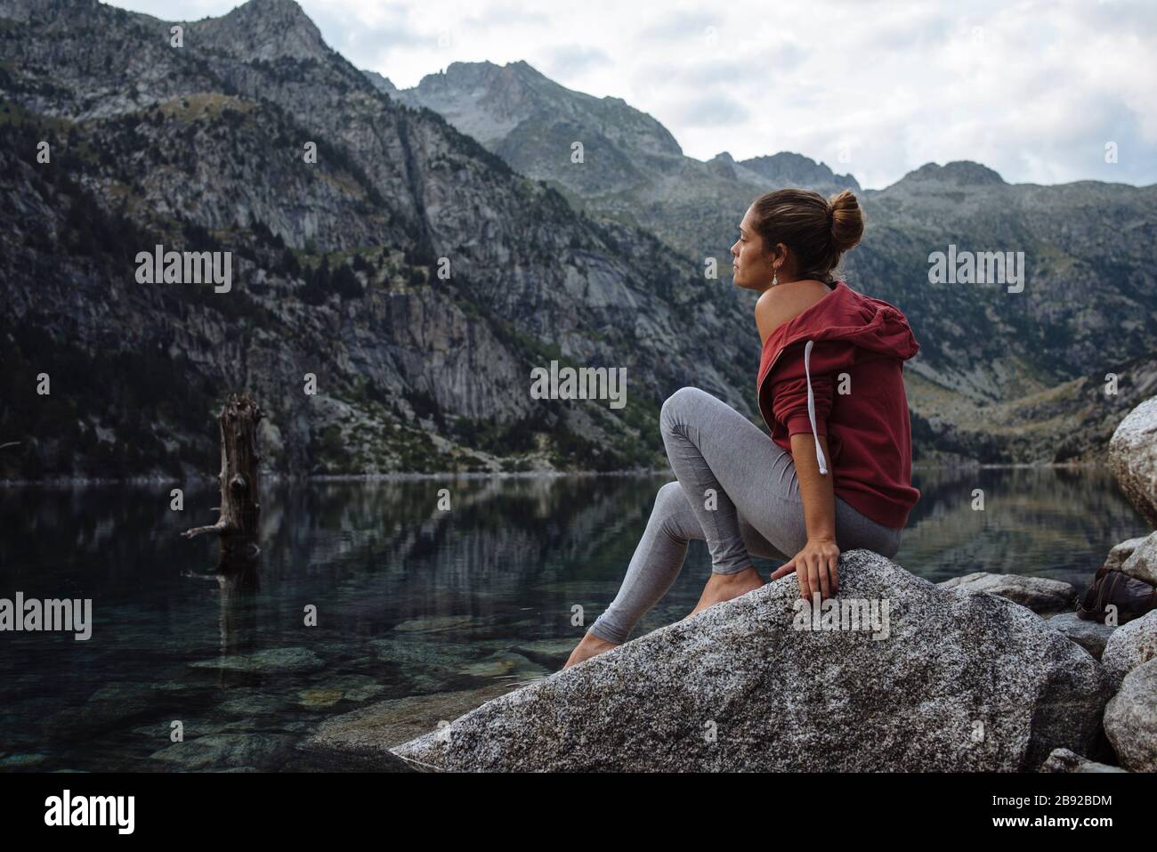 Woman with bun sitting on a rock by a lake during a trip. Stock Photo