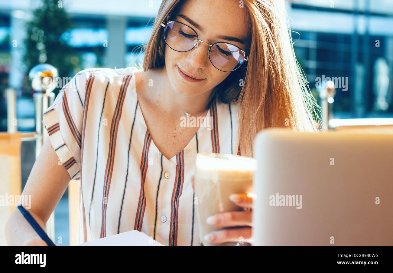 Ginger business lady with eyeglasses is writing something while using a laptop and drinking a cocktail at the coffee shop Stock Photo