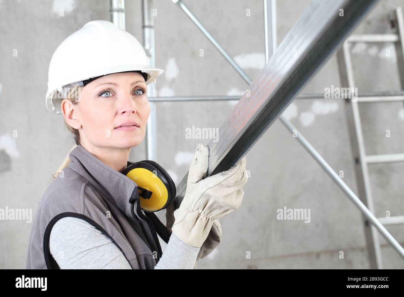 woman construction worker builder portrait wearing white helmet and hearing protection headphones, holding a metal stud for drywall on interior site b Stock Photo