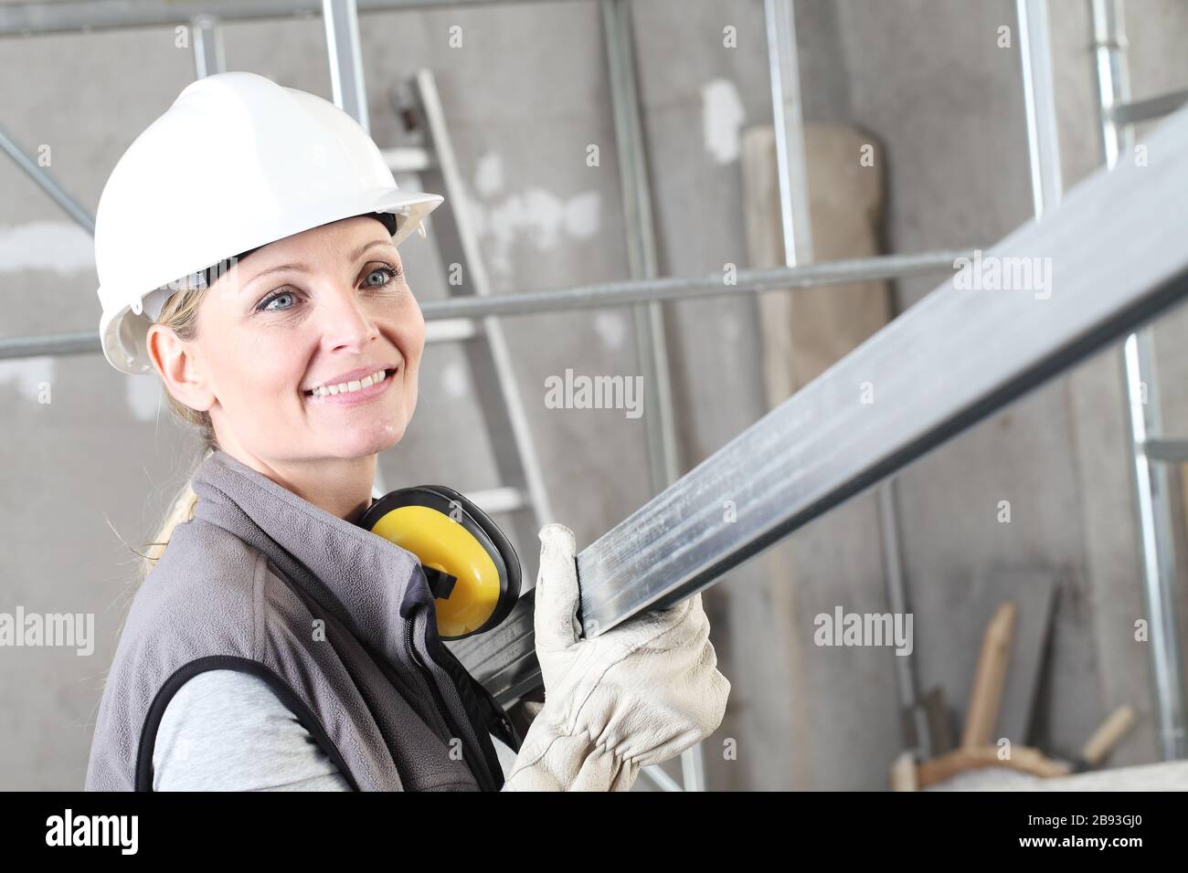 smiling woman construction worker builder portrait wearing white helmet and hearing protection headphones, holding a metal stud for drywall on interio Stock Photo