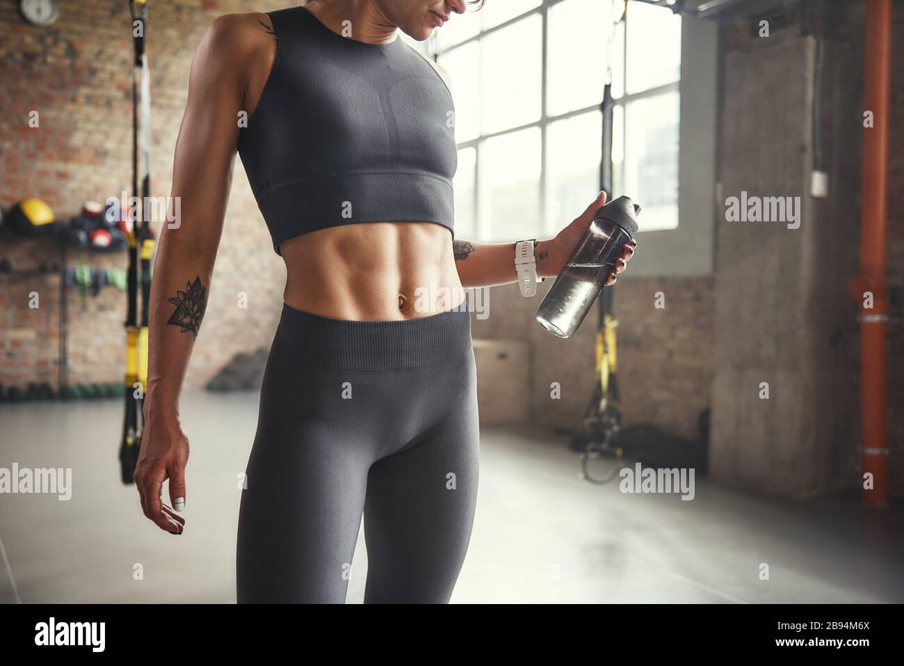 Healthy lifestyle. Cropped photo of young athletic woman in sports clothing holding bottle of water while exercising in gym. Professional sport. TRX Training. Stock Photo