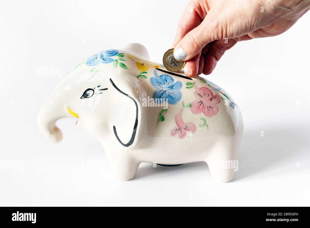 Female hand with silver nail varnish inserting a pound coin into a ceramic elephant money box Stock Photo