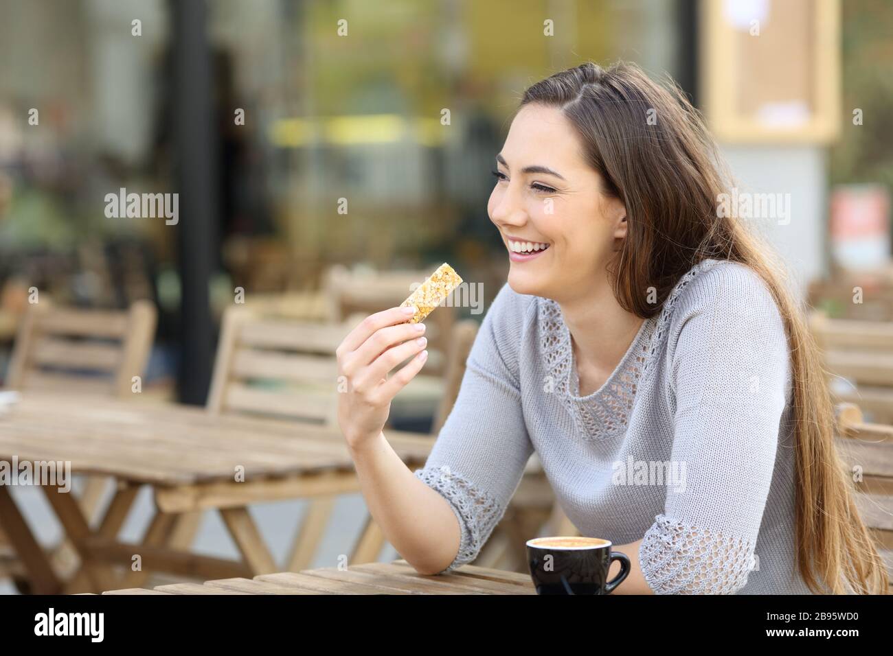 Happy young woman holding a cereal snack bar sitting on a cafe terrace Stock Photo