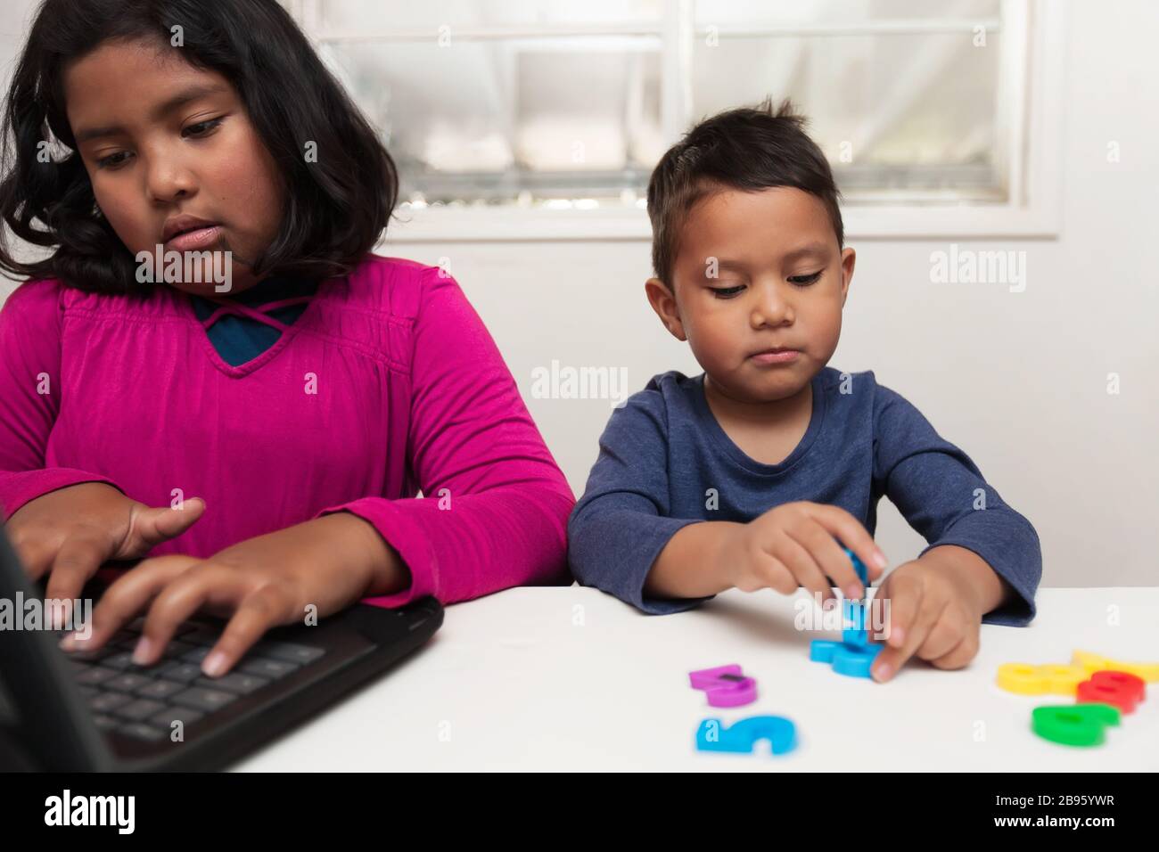 Children in home based learning, with the older sister typing on a laptop while her little brother plays with colorful number manipulatives. Stock Photo
