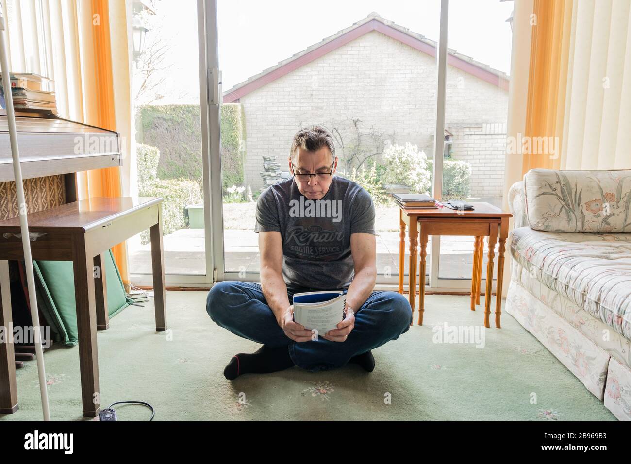 50 something male in coronavirus self isolation at home, sitting in front of window in living room, reading a book Stock Photo