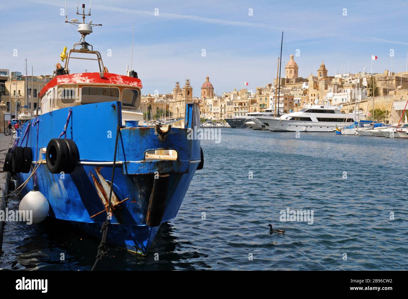 A fishing boat at the dock in Cospicua, Malta, one of the Three Cities, with luxury yachts and the city of Birgu/Vittoriosa in the background. Stock Photo