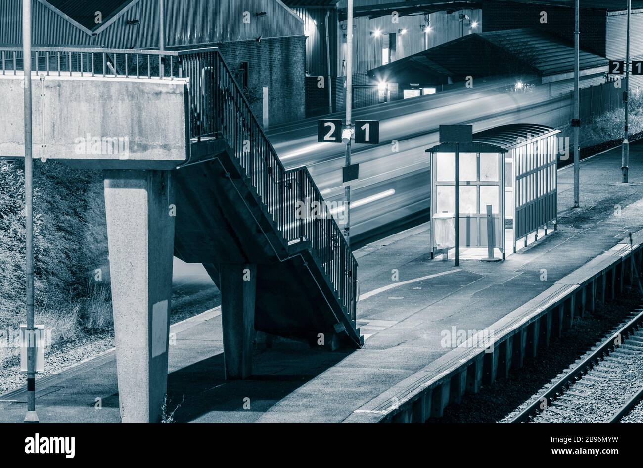 Speeding train passing through empty suburban railway station (with no wheelchair access) at night. UK Stock Photo