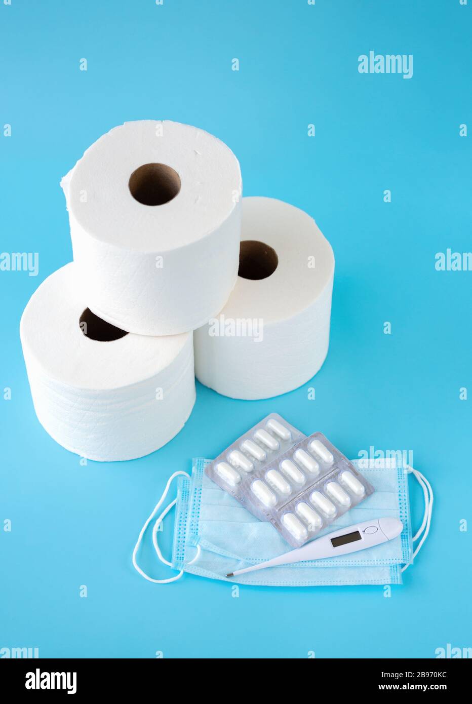 Face mask, thermometer, pills, tree toilet paper rolls on blue background. Medicine supplies for home coronavirus COVID-19 quarantine. Copy Space Stock Photo