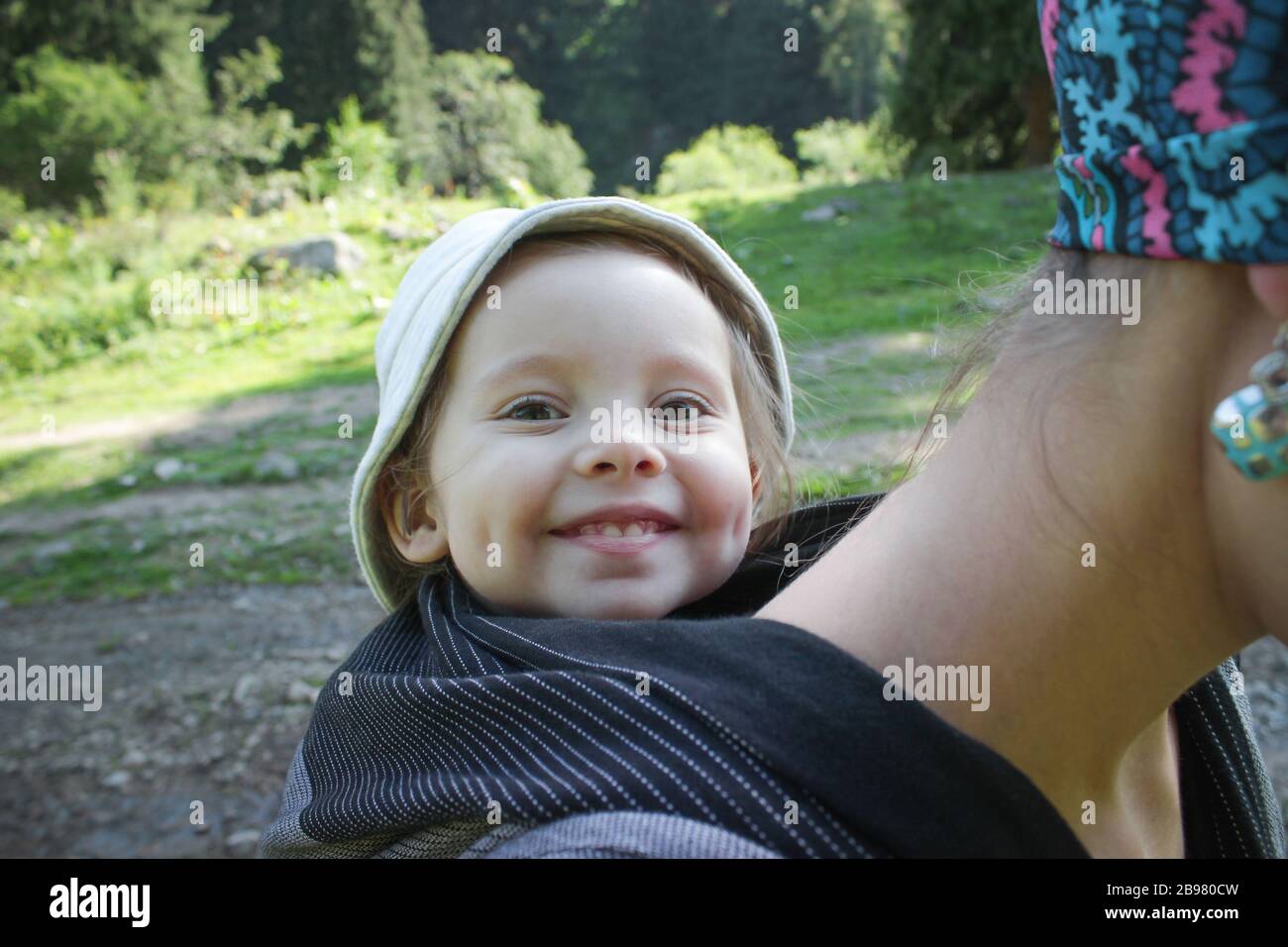 Mother carrying a two year old smiling baby in a black woven wrap. Tracking, outdoor Stock Photo