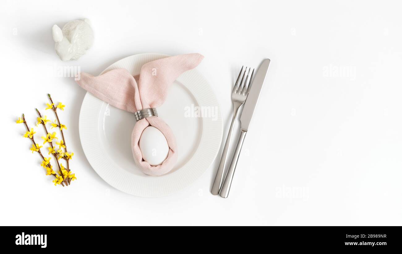 Easter holiday table setting with egg in pink napkin Easter Bunny and yellow forsythia flowers on a white background. Copy space, top view, flat lay. Stock Photo