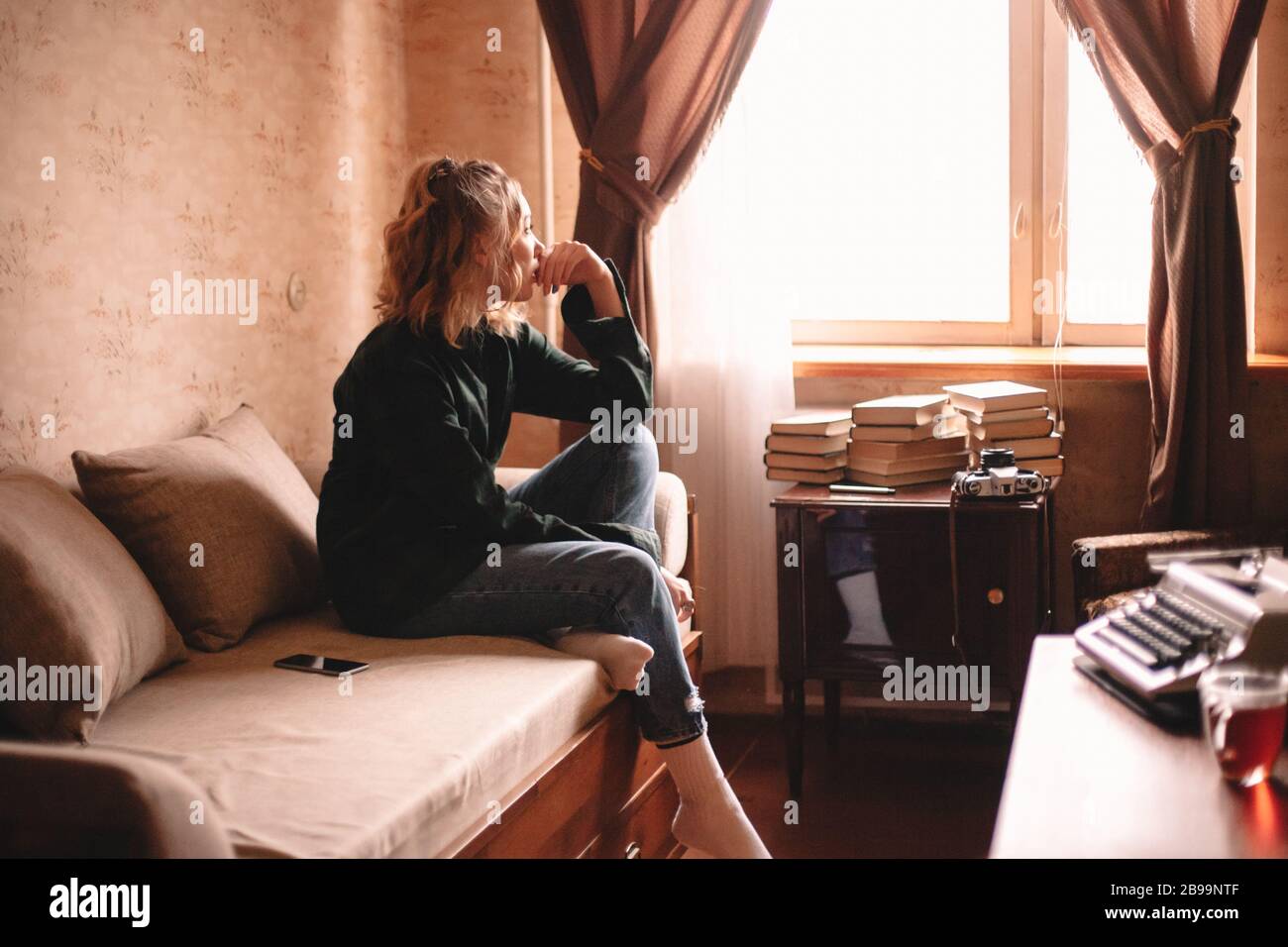 Thoughtful young woman looking through window while sitting on bed at home Stock Photo
