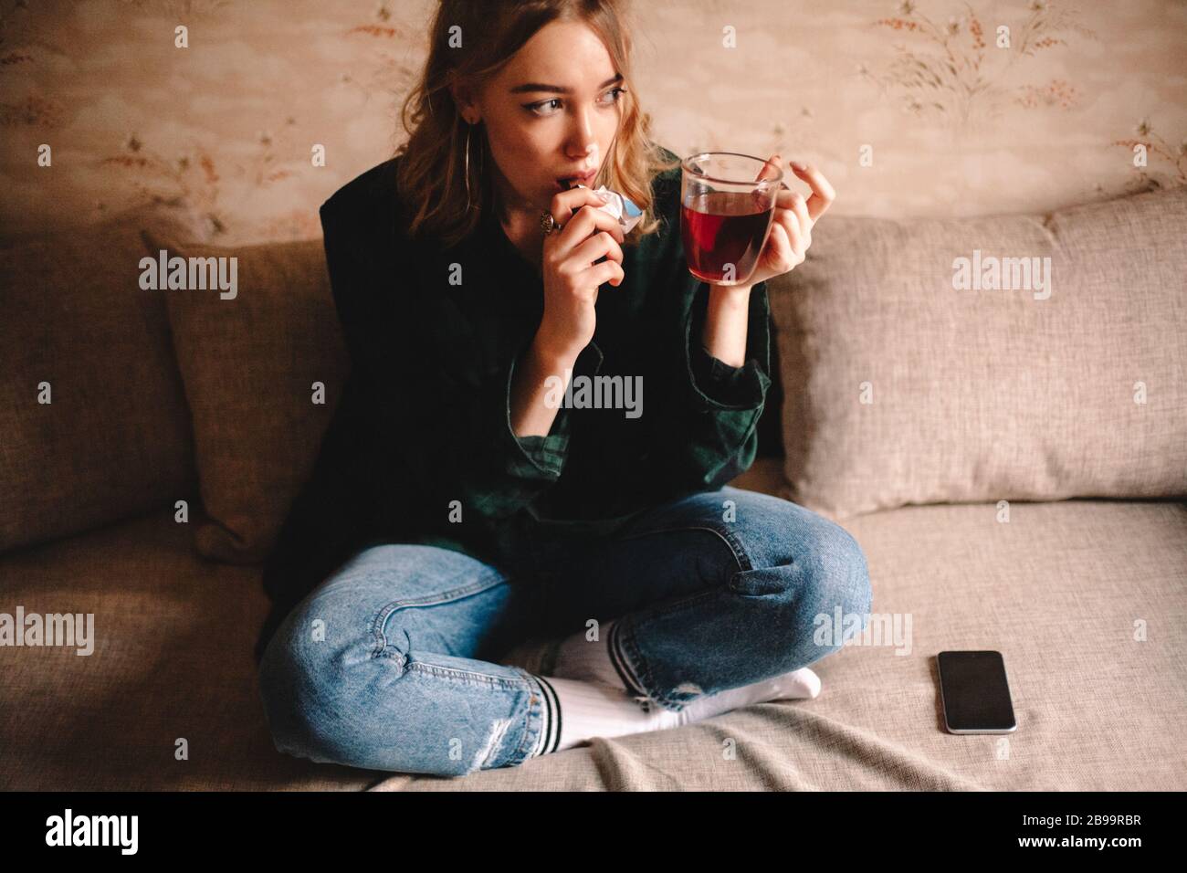 Young thoughtful woman drinking tea and eating chocolate while sitting on sofa at home Stock Photo