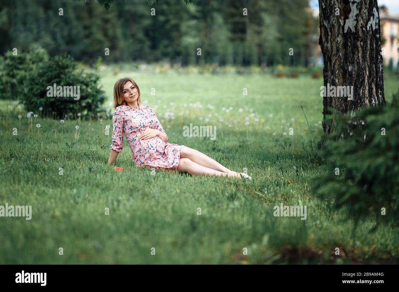 Pregnant woman inpink dress sitting on grass and touching bump whilst holding pink rose girl waiting Stock Photo