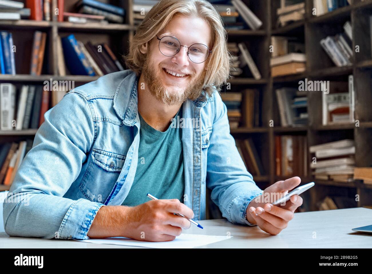 Portrait of happy smiling young student at library desk hold modern smartphone. Stock Photo