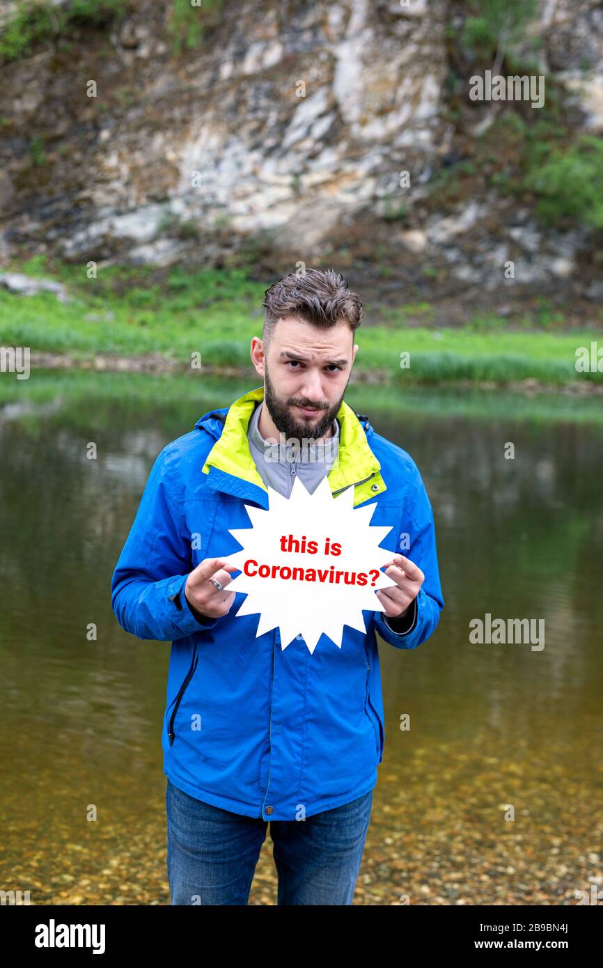 Portrait of handsome young man in jeans and blue sport jacket on bank of clear mountain lake with cartoon star plate asking if this is coronavirus in Stock Photo