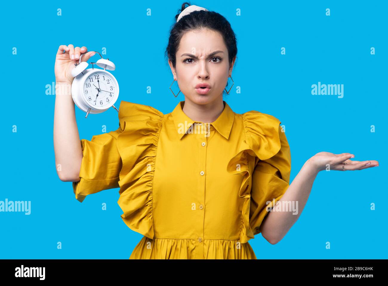 Woman in a mustard dress holding a clock and looking uncertain Stock Photo