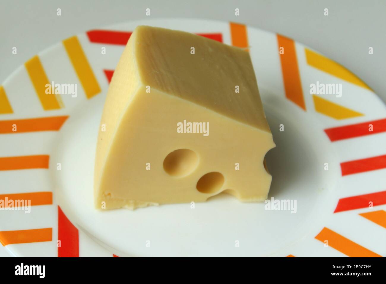A piece of yellow cheese with holes lies on a white plate with a colored edge. Stock Photo
