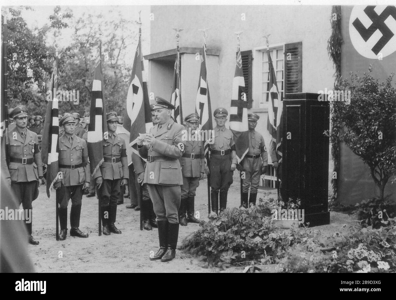 A senior official of the NSDAP gives a speech in Ochsenfurt and reads it from his notepad. Other members of the NSDAP with swastika flags stand in a line. [automated translation] Stock Photo