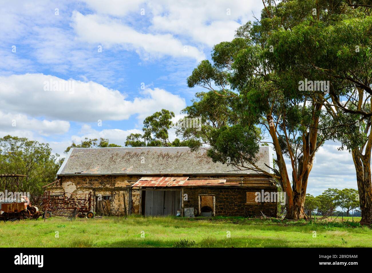 Old Stone Farmhouse in Disrepair Under Tree with Old Machinery Stock Photo