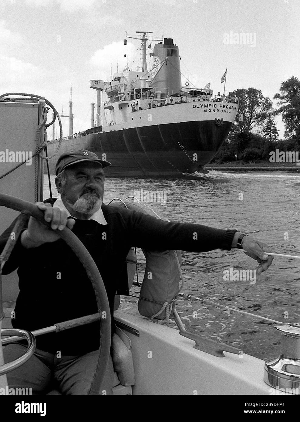 A man with beard and cap steers a boat through the Kiel Canal. In the background you can see a cargo ship. [automated translation] Stock Photo