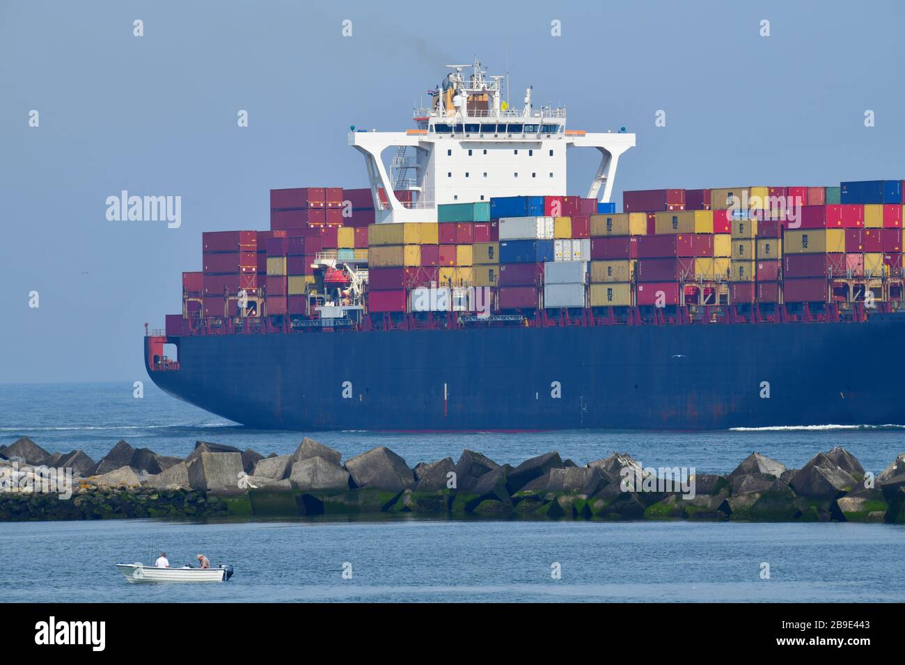 Arrival of large containership in Rotterdam harbor with in the forefront a tiny boat with 2 man fishing that dwarfs in comparison Stock Photo
