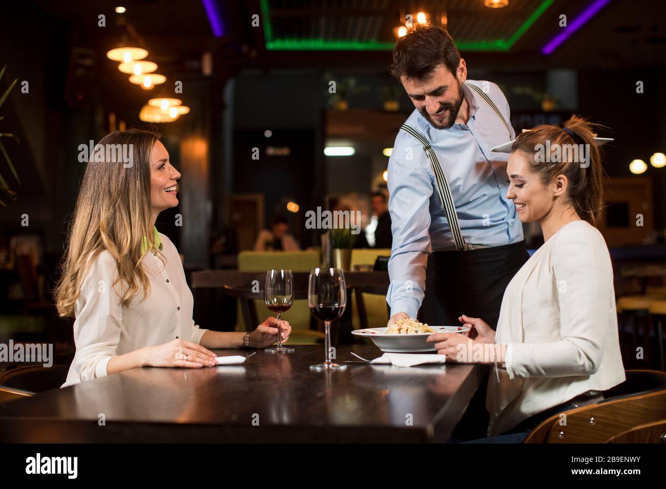 Two smiling young female friends at a restaurant with waiter serving dinner  Stock Photo - Alamy