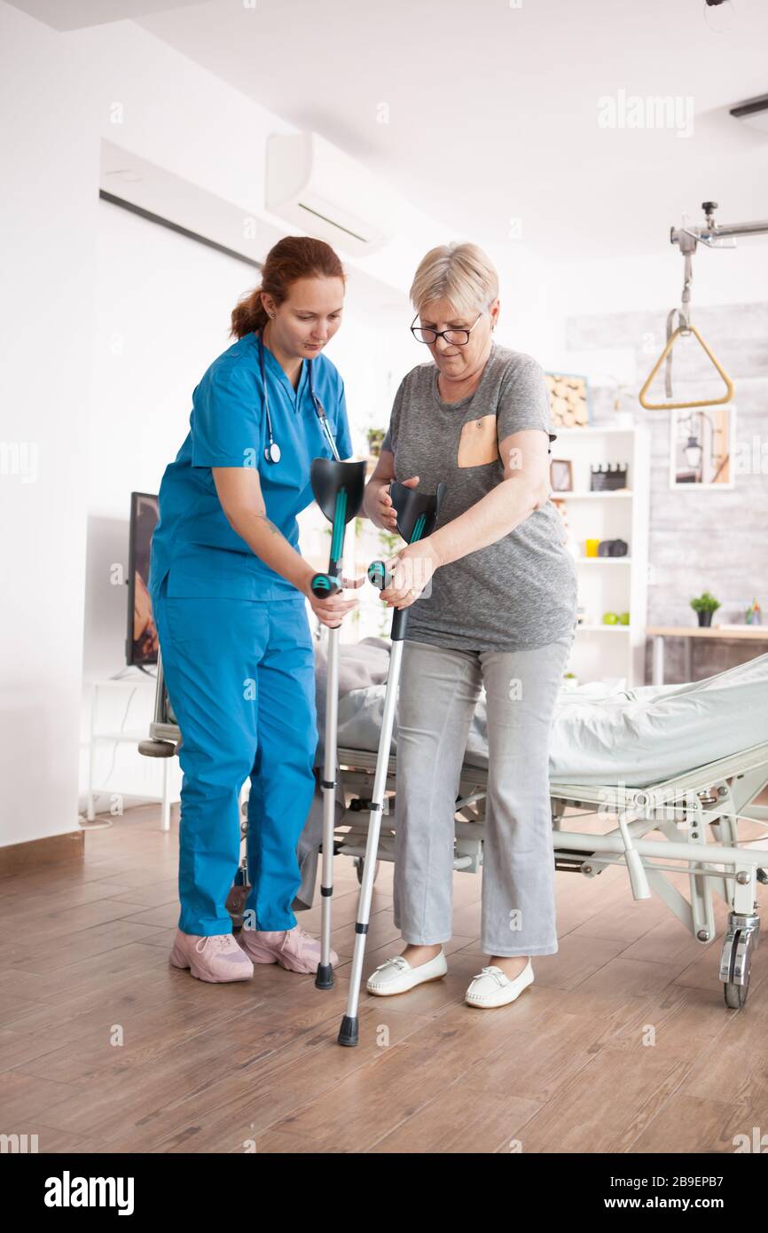 Female caregiver helping elderly age woman in nursing home to walk with