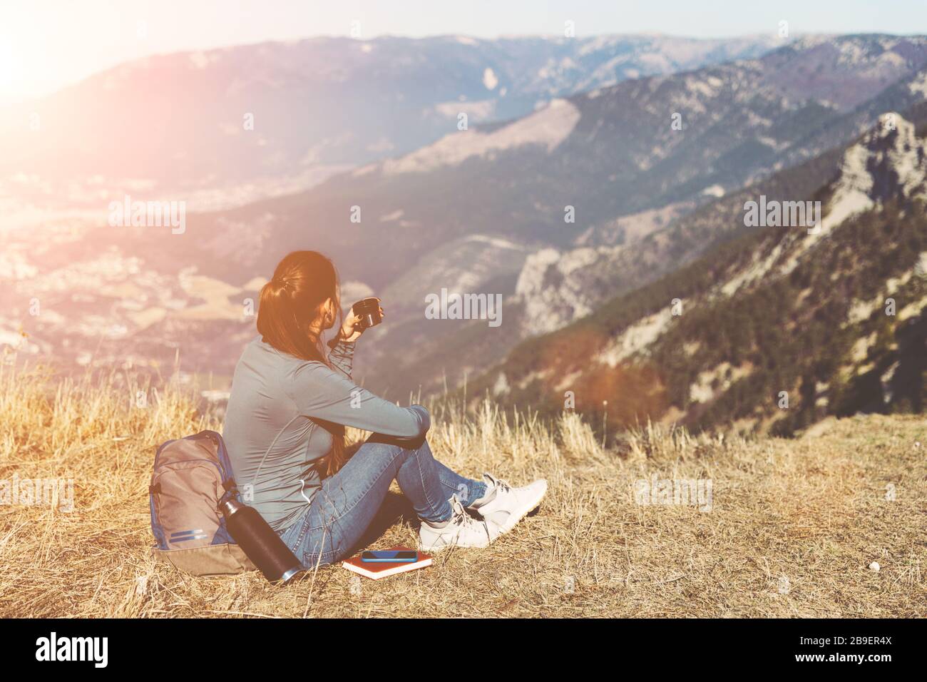 Young beautiful girl travels alone in the mountains in spring or autumn, sits on the edge of the mountain and looks into the distance and enjoys nature, rocks and green forests, view of the landscape. a backpack behind and sportswear, a thermos with a hot drink or tea, freedom and lightness. Back view of woman traveler in cap sitting on mountain top alone and looking at beautiful summer landscape and blue sea view. Drink tea or coffe Stock Photo