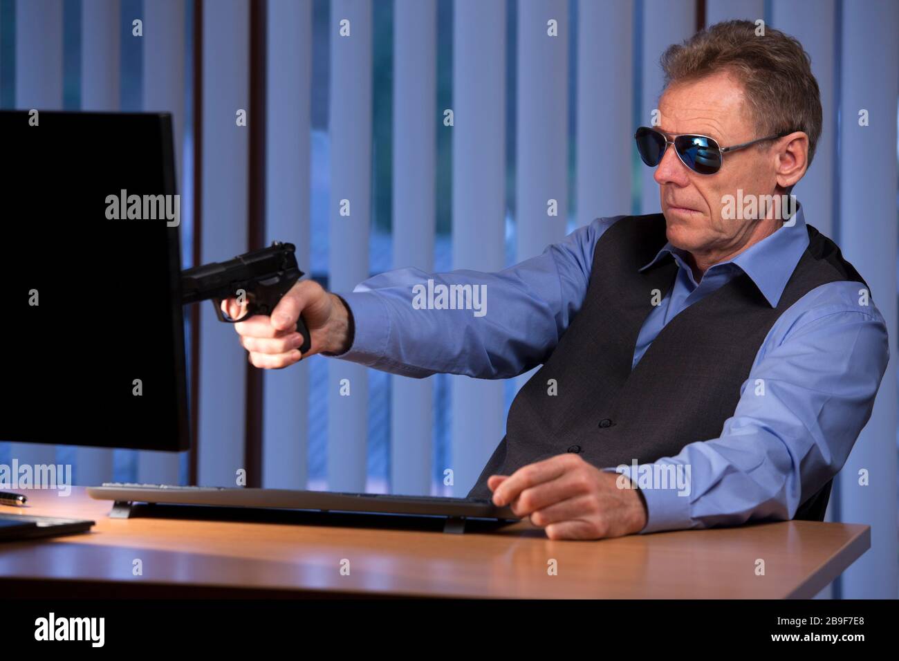 Criminal businessman sitting at a desk aiming with pistol at a computer monitor in a dark office by night Stock Photo