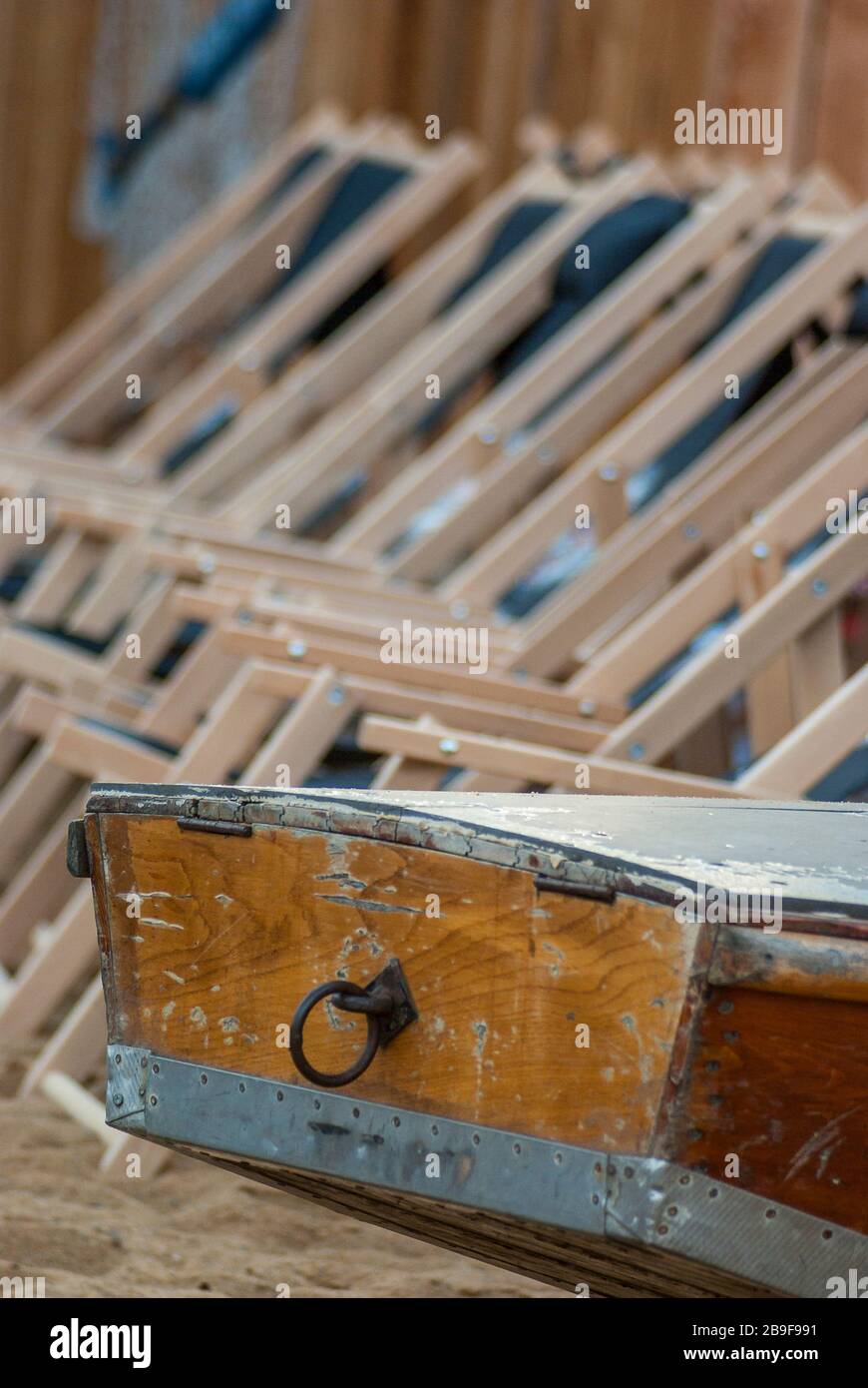 the boat tail of a wooden rowing boat and in the background a series of suspended empty deckchairs lined up in order on the beach in the sand, das Hec Stock Photo