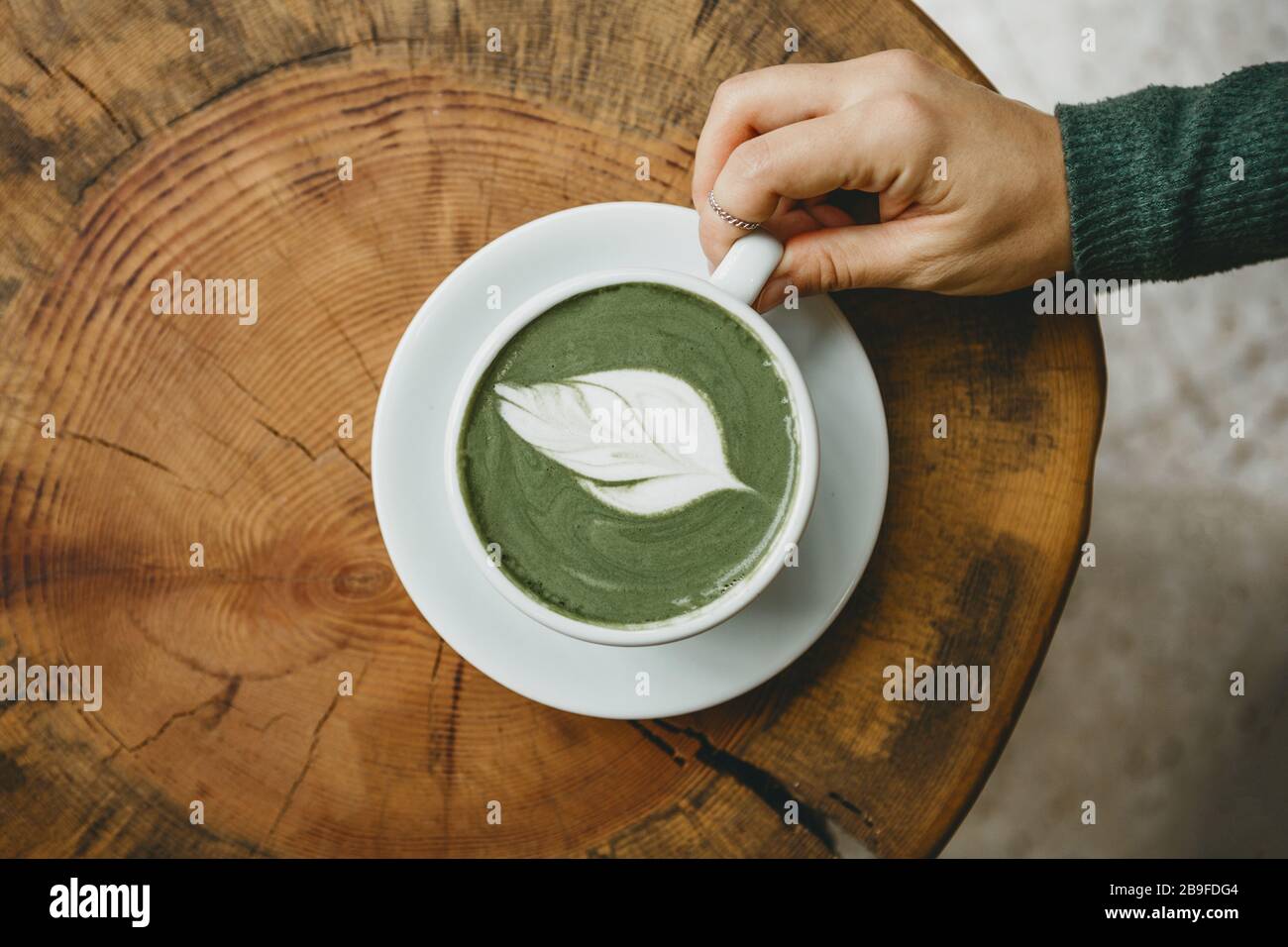 View from above. Girl holds in her hand a cup with fragrant fresh and healthy green matcha latte tea on a wooden table. Stock Photo