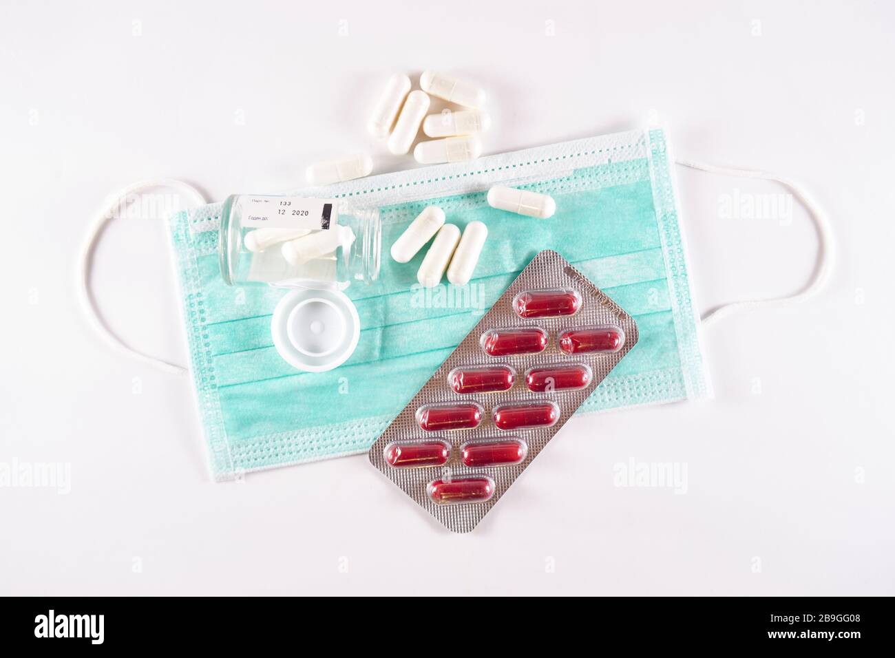 White medicine capsules spilling out of glass bottle next to a blister with red pills on top of a surgical mask (top view on white background) Stock Photo