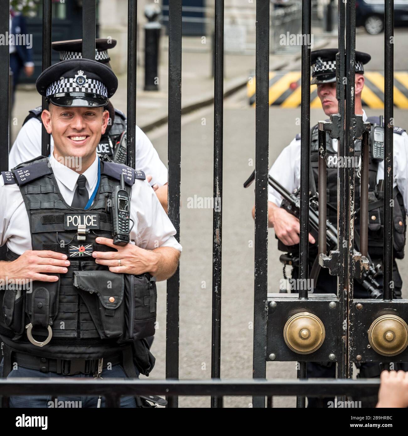 Armed Guards. A friendly London policeman standing guard outside the gates of Downing Street with armed colleagues in the background. Stock Photo