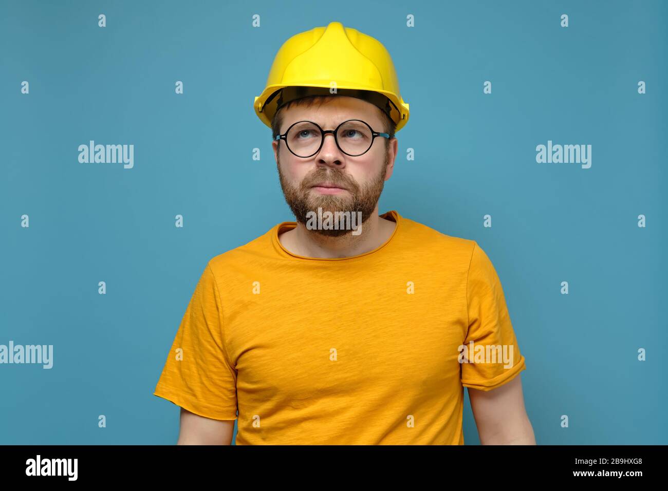 Puzzled bearded man in a construction helmet and round glasses looks up thoughtfully, on a blue background. Stock Photo