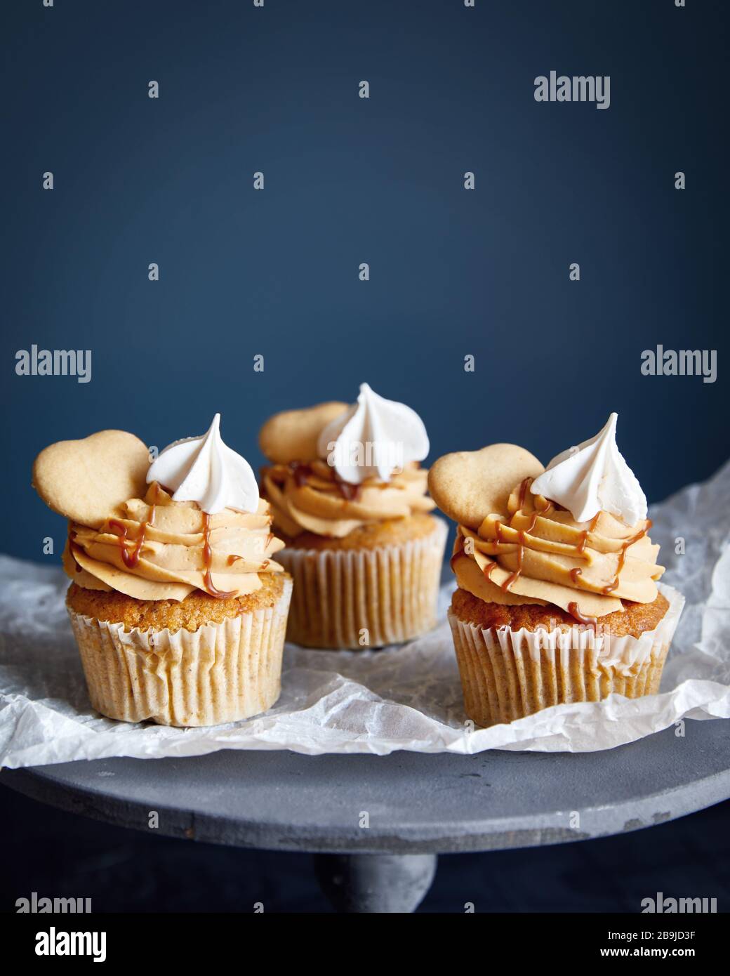 Carrot cupcakes with walnuts and spices decorated with heart shape biscuit, meringue and salty caramel on white paper and dark background. Free space Stock Photo