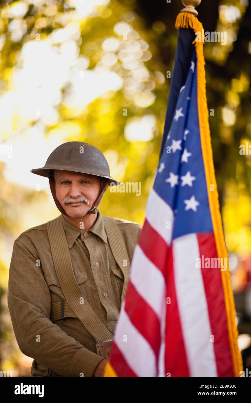 Mature male reenactor wearing an American World War steel combat helmet and uniform and holding up American flag poses for a portrait. Stock Photo