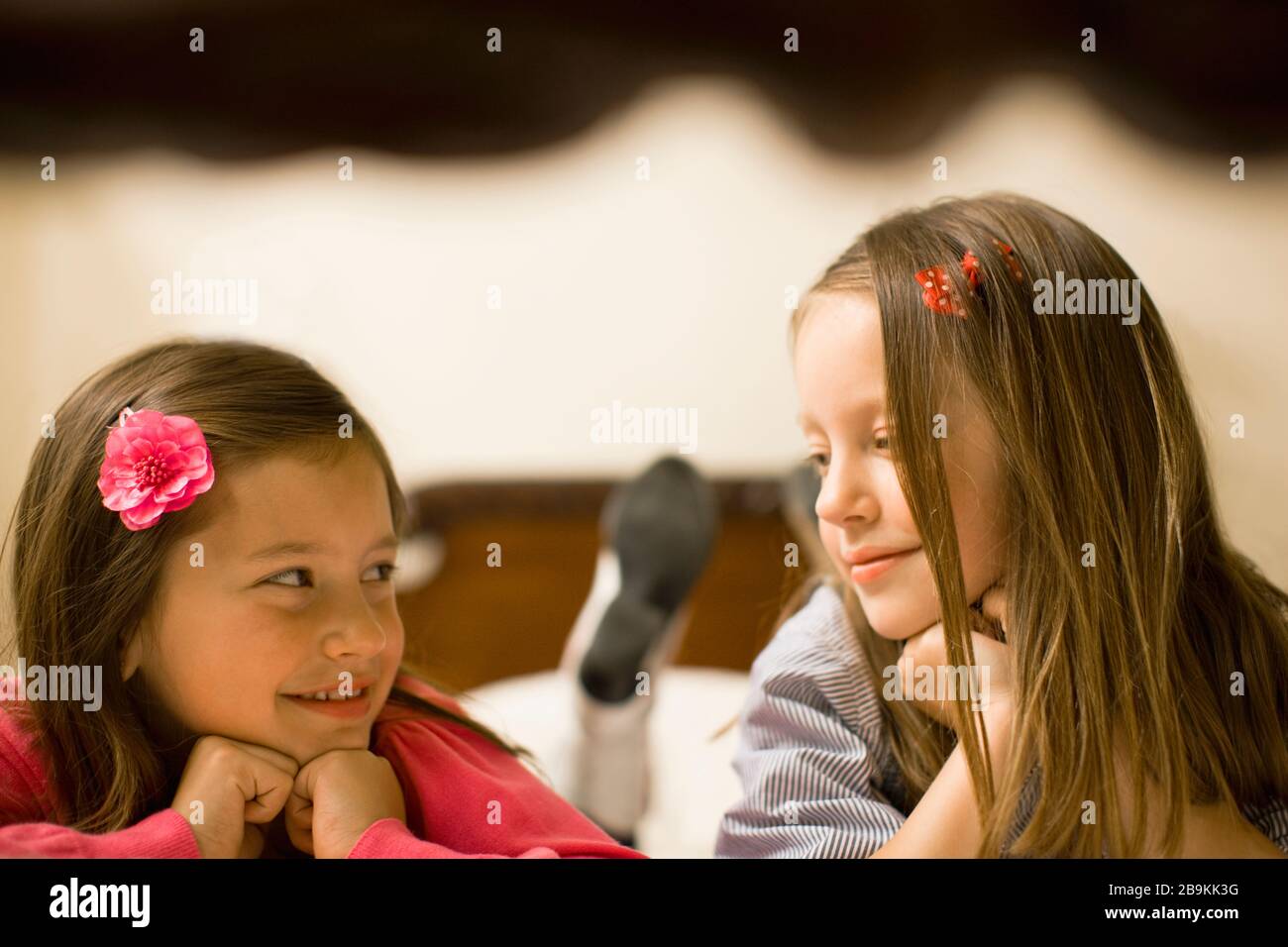 Two young girls smile at each other as they lie front down on a bed
