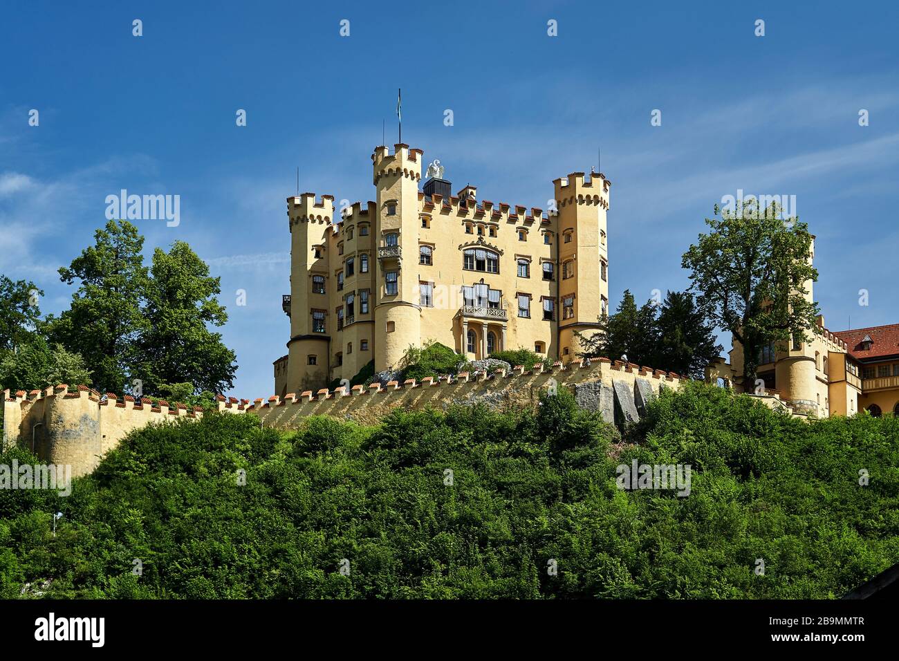 A view of a Hoehenschwangau castle on top of a hille on a sunny summer day against a blue sky and green foliage bellow the castle wall. Stock Photo