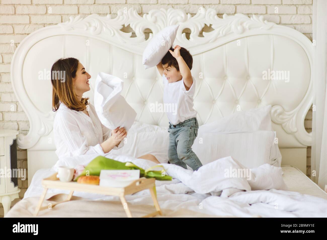 Beautiful mother playing pillow fight with her cute son in bedroom in the morning. Stock Photo