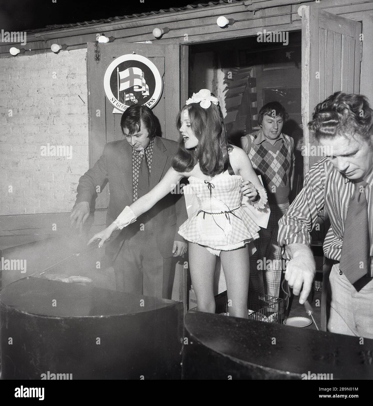 1973, historical, attractive young lady wearing a very short and revealing party frock helping with the cooking outside on a grill at a fund raising barbeque at The Lord Northbrook pub, Lee, South East London, England. At the corner of southbrook Rd, the pub is named after Lord Northbrook, whose family is Baring, the banking family who in the 19th century had an estate in the borough. Stock Photo
