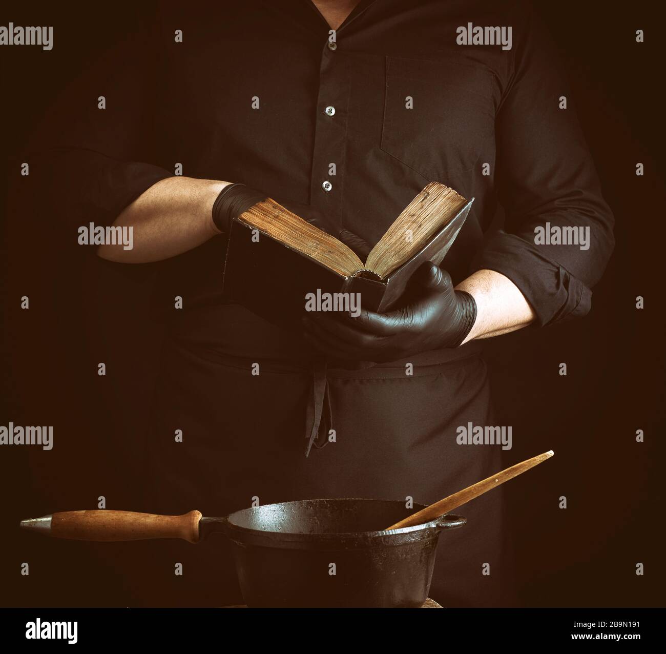 male cook in a black uniform holds an old cookbook with recipes, on a table is a cast-iron frying pan with a wooden handle, low key Stock Photo