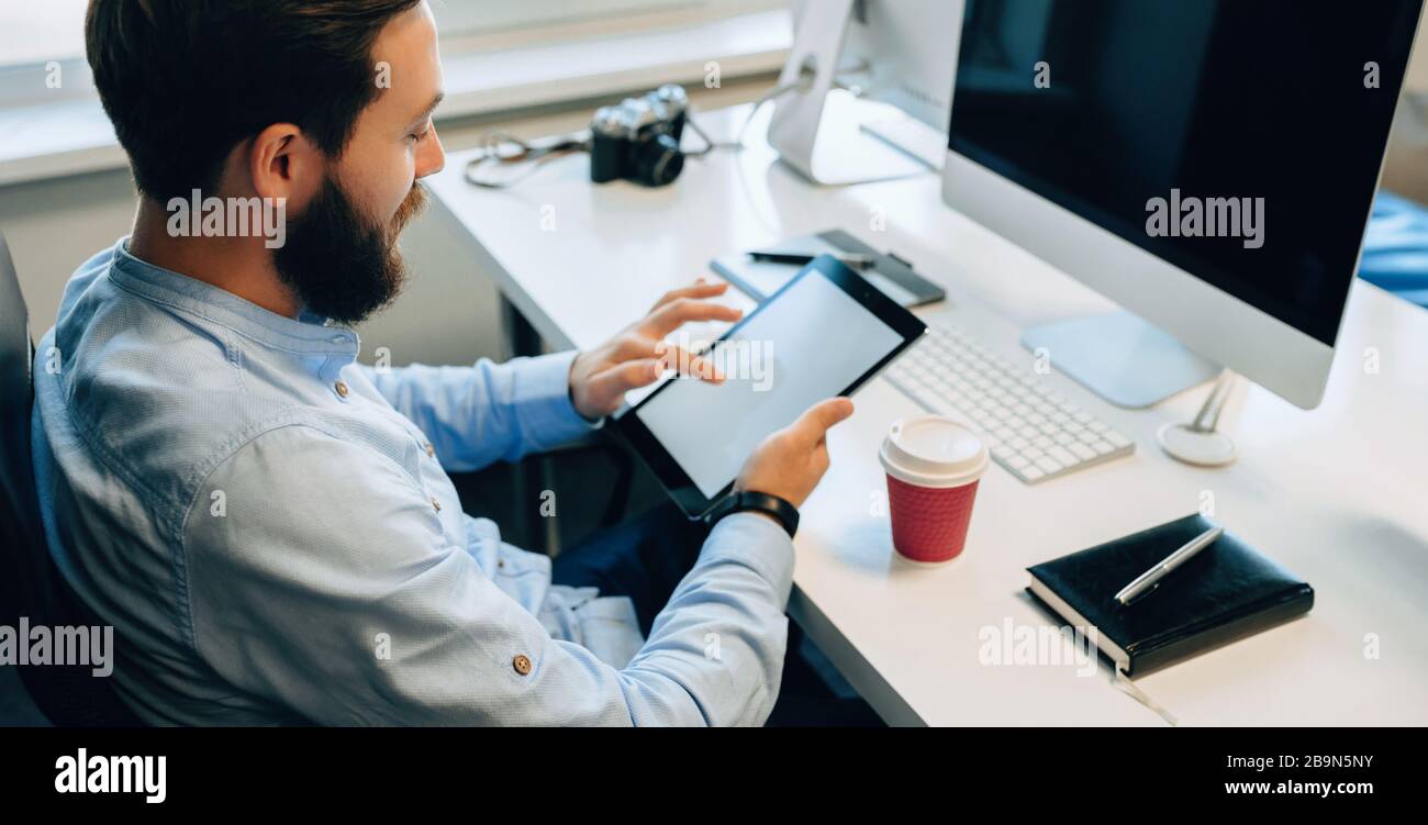 Upper view portrait of a bearded caucasian man using a tablet with blank space while drinking a coffee in his office Stock Photo