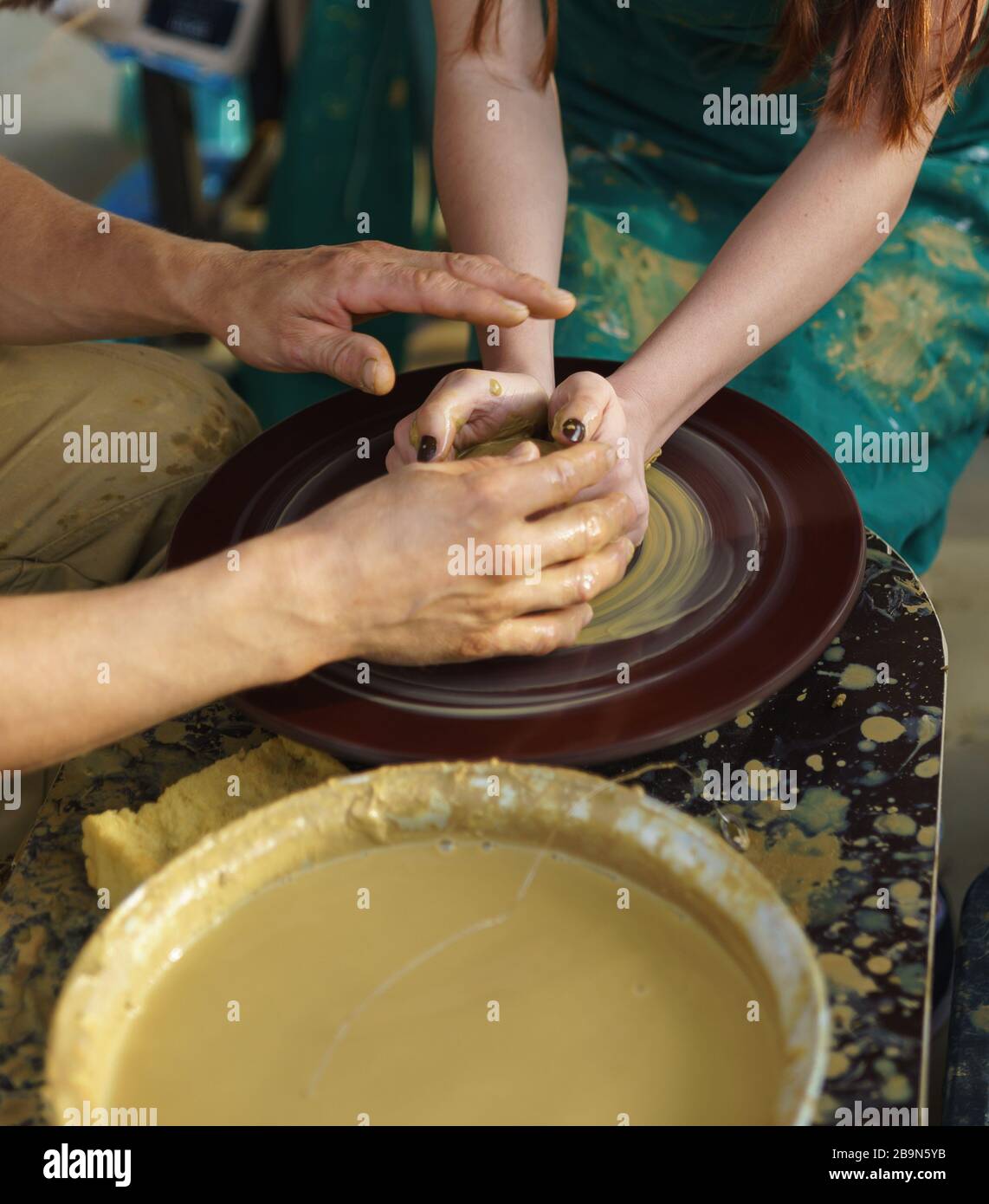 Male hands of the master and the hands of a girl in a green apron on a Potters wheel make a pitcher of yellow clay Stock Photo