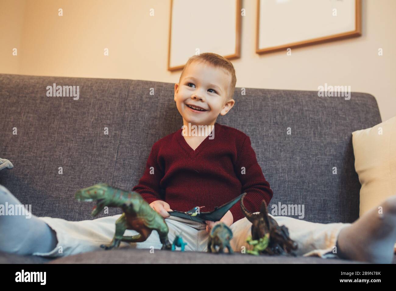 Caucasian small boy sitting on the sofa and playing with his dinosaur toys while looking at his parents Stock Photo