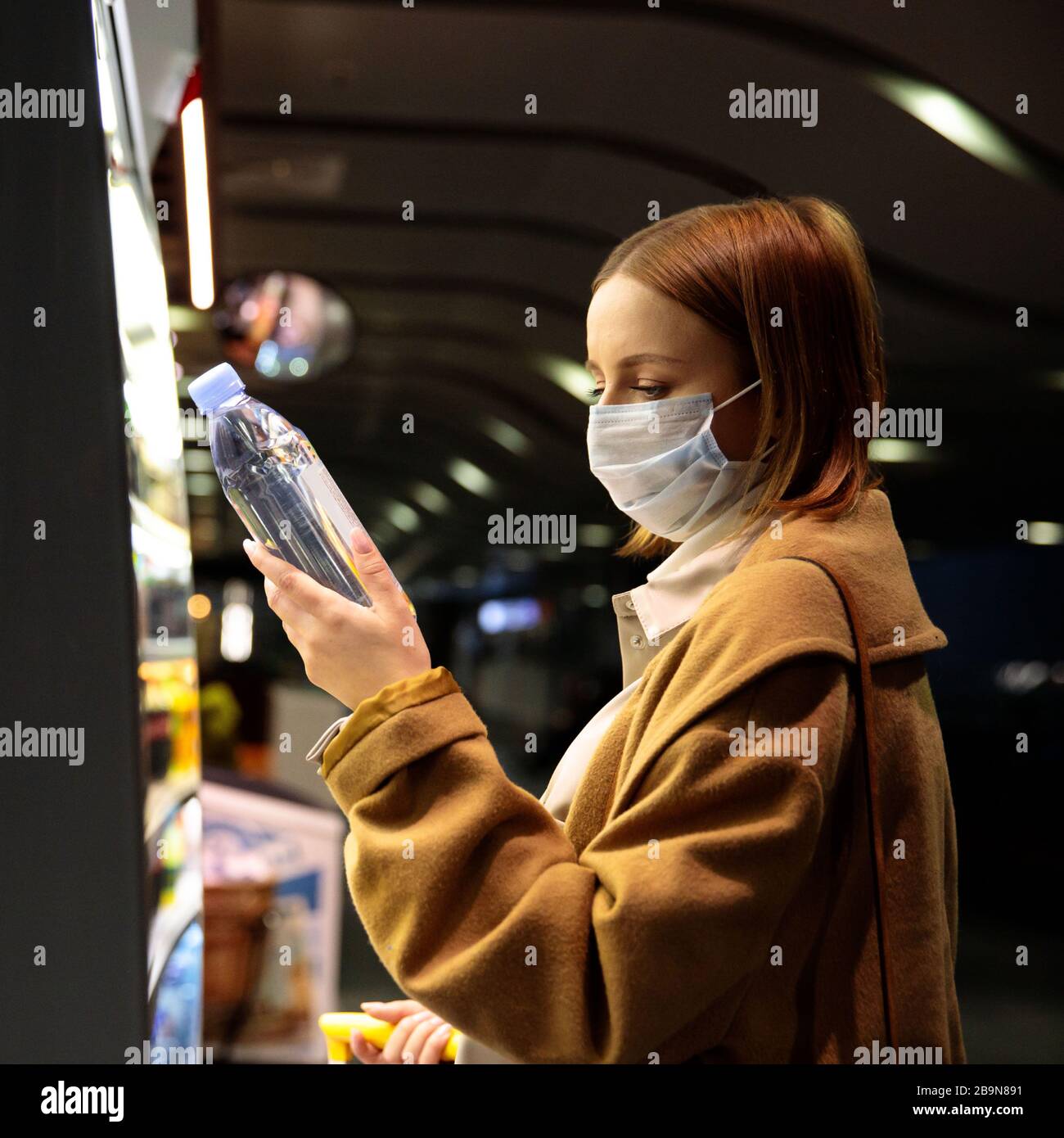 Caucasian woman customer wearing facial protective mask in airport, buying and choosing water bottle, reads information on a label. Purchase of food d Stock Photo