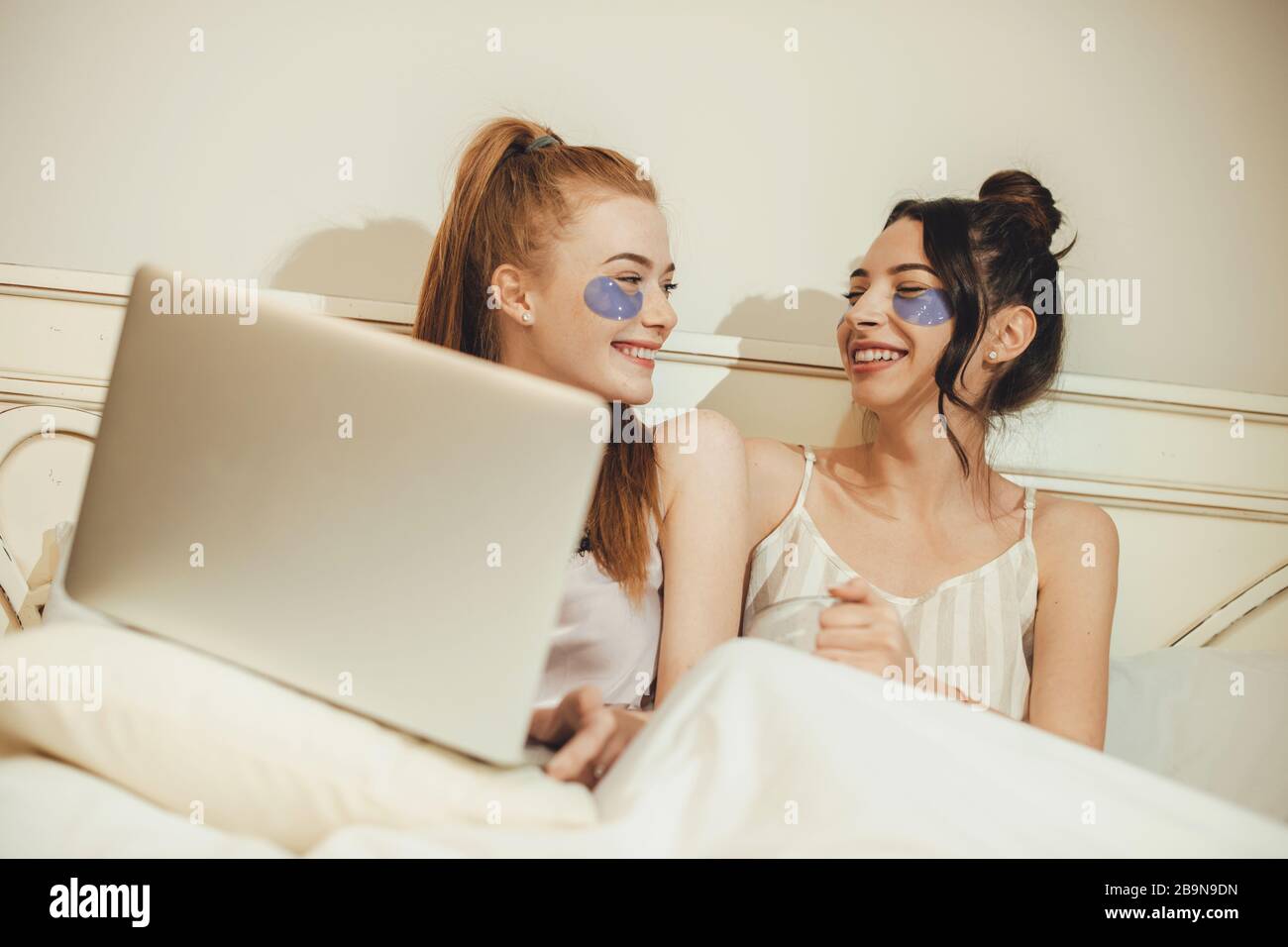 Two caucasian sisters wearing some anti aging masks while lying in the bed with a computer Stock Photo
