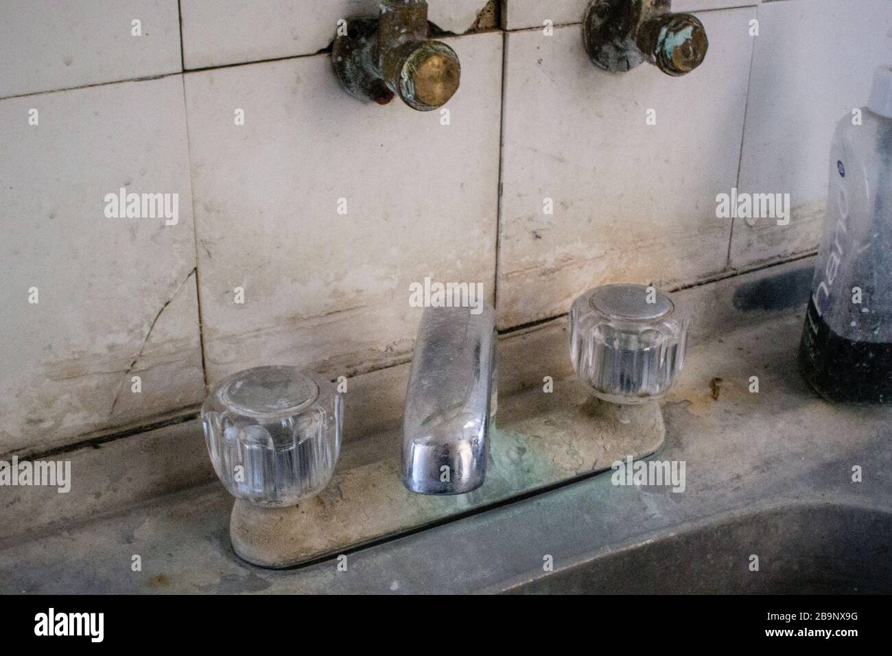 Stainless steel kitchen sink with heavily stained, oxidised and encrusted mixer tap, dirty and cracked white tile splash back and hand wash bottle Stock Photo