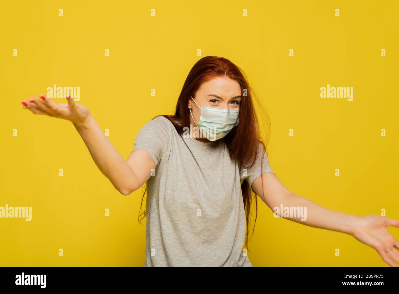 Beautiful caucasian young woman in gray t-shirt with disposable face mask. Protection versus viruses and infectious. Studio portrait, concept with yellow background. Shocked woman waving her arms. Stock Photo