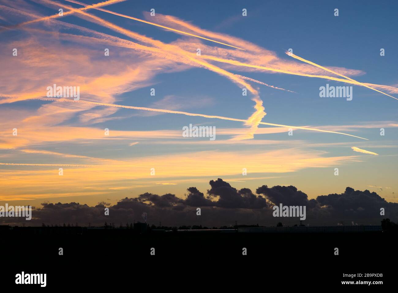 Colorful contrails high up in the sky are illuminated by the light of the setting sun. Closer to the surface of the earth, there are dark clouds. Stock Photo