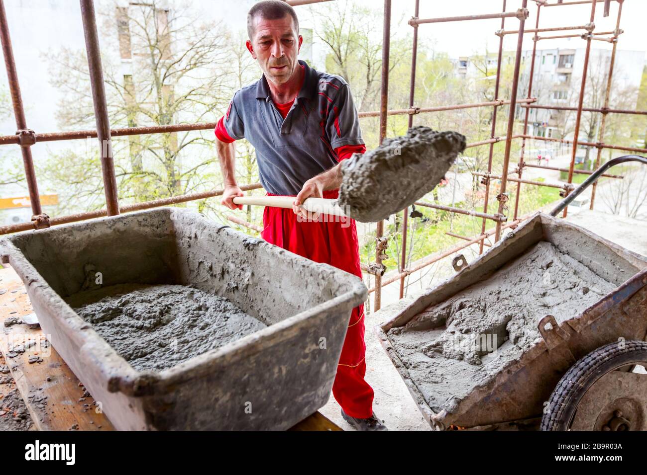 Construction worker is using shovel to unloading cement, mortar, from wheelbarrow on construction site. Stock Photo